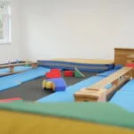 Blick in unsere Turnhalle in Schwerte. Spielgeräte liegen im Raum herum. Es sind verschiedenen Turngeräte zu einem Parcours aufgebaut. Um den Parcours herum liegen viele blaue Turnmatten.